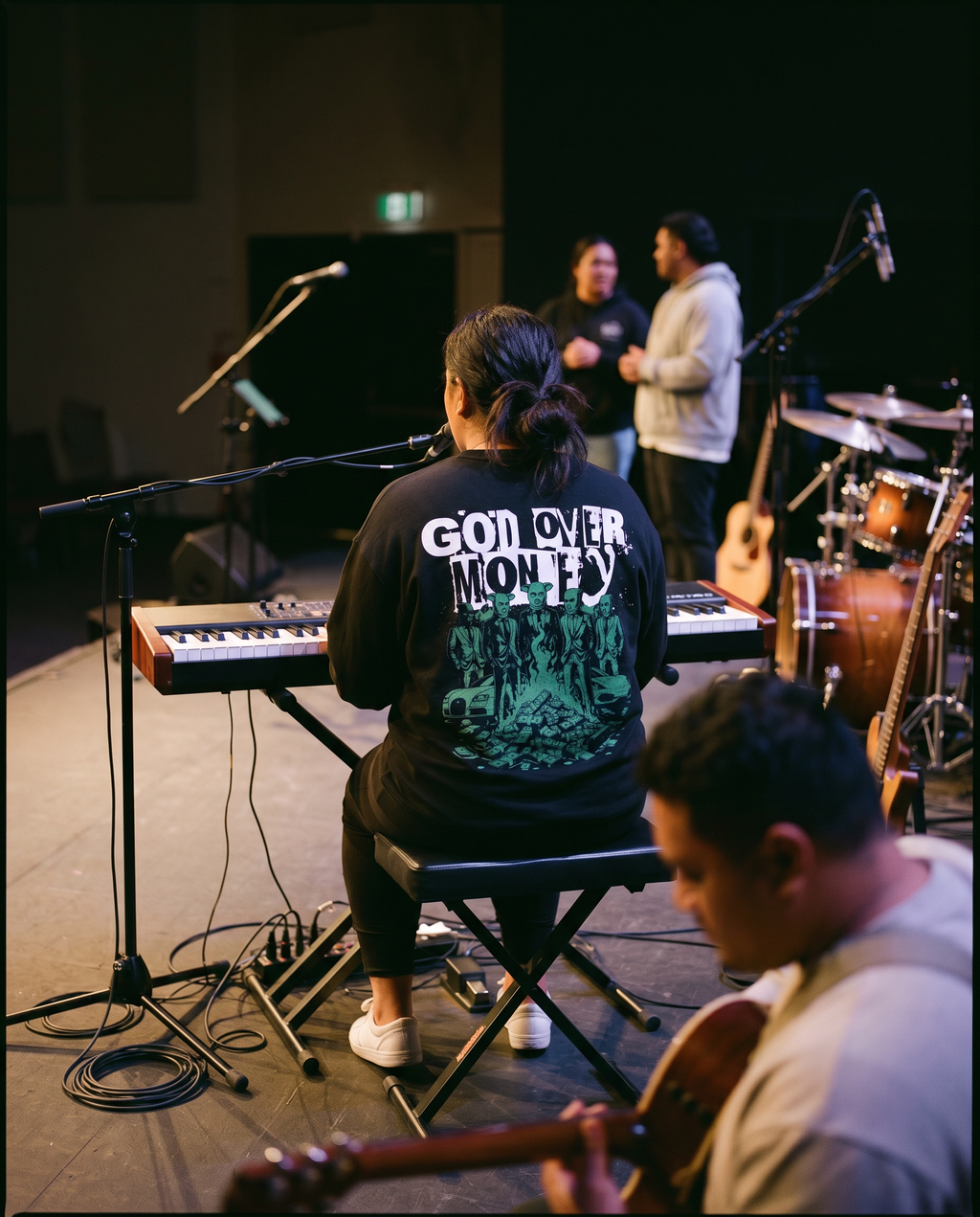 Musician playing keyboard with 'God Over Money' shirt, another person with guitar, and a third person in the background.